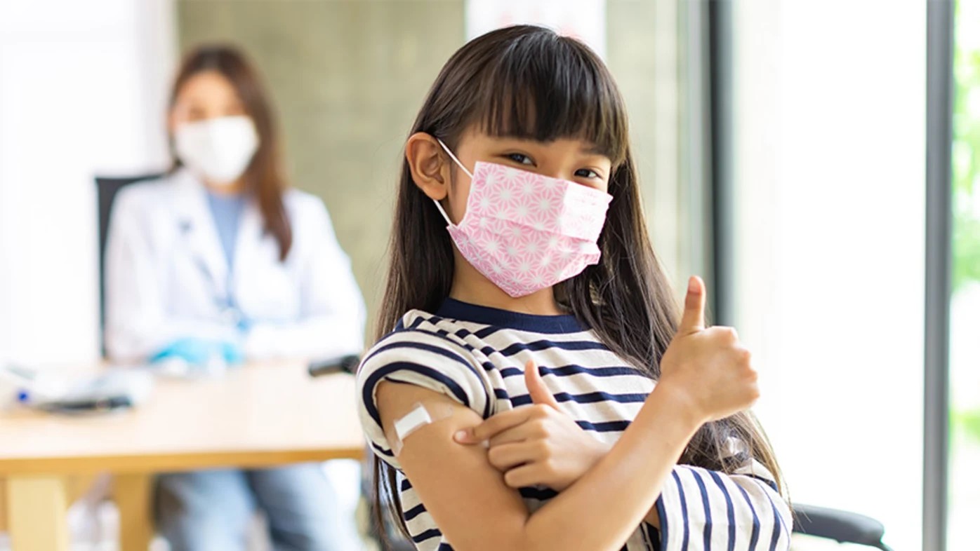 Niña pequeña con mascarilla levantando los pulgares después de recibir una vacuna